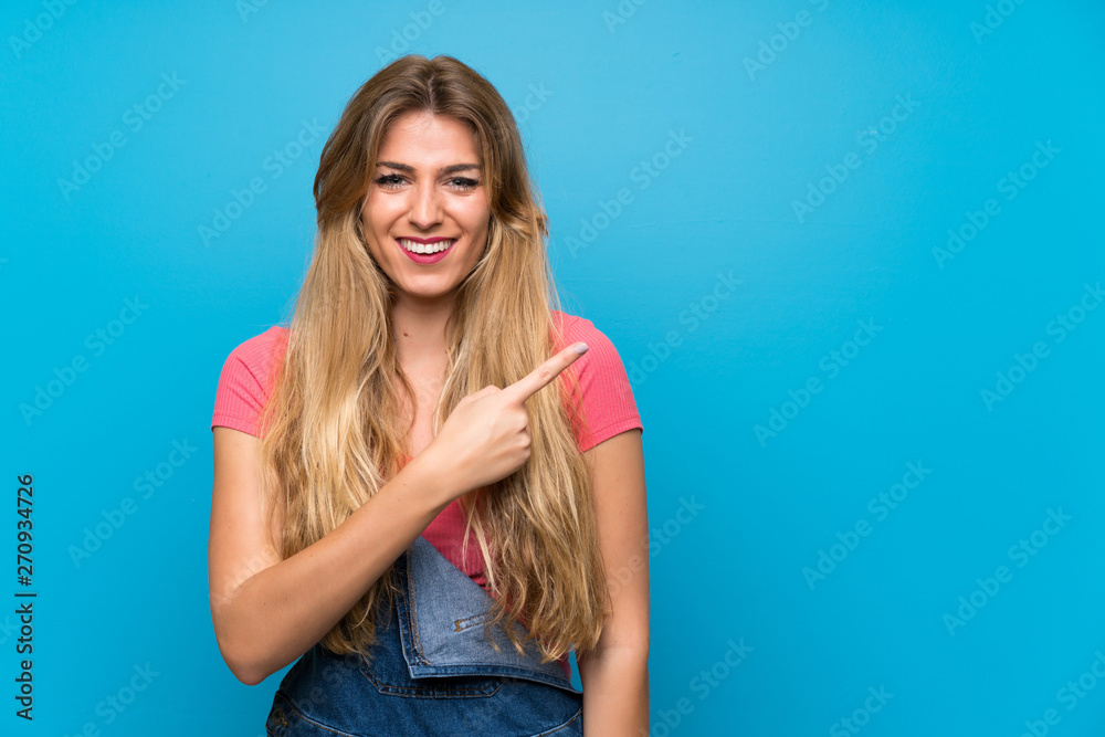 Young blonde woman with overalls over isolated blue wall pointing to the side to present a product Young blonde woman with overalls over isolated blue wall pointing to the side to present a product