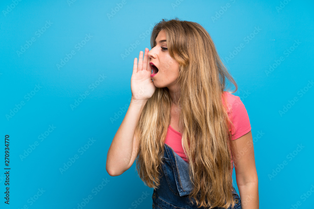 Young blonde woman with overalls over isolated blue wall shouting with mouth wide open to the lateral