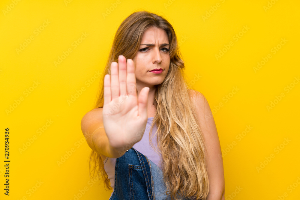 Young blonde woman with overalls over isolated yellow background making stop gesture with her hand Young blonde woman with overalls over isolated yellow background making stop gesture with her hand