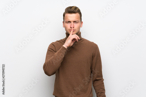 Blonde man over isolated white wall showing a sign of silence gesture putting finger in mouth