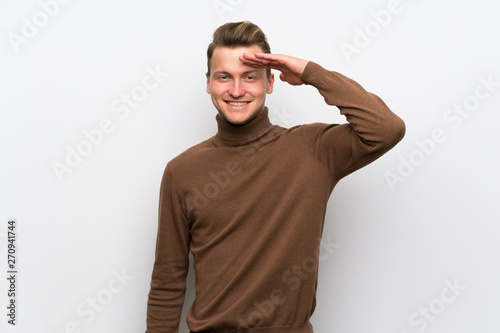 Blonde man over isolated white wall saluting with hand