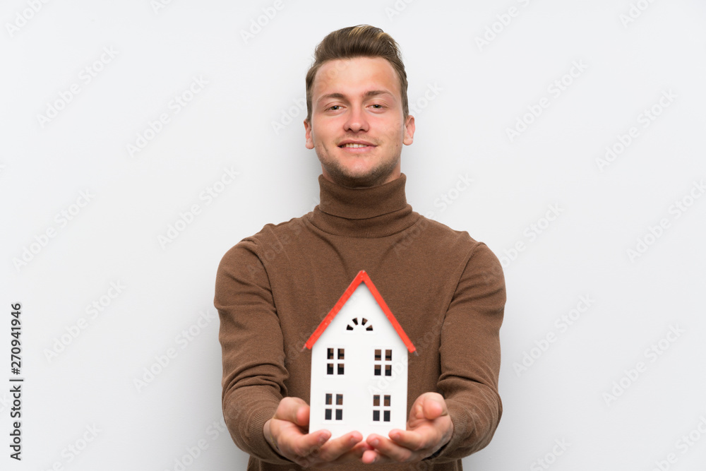 Blonde man over isolated white wall holding a little house