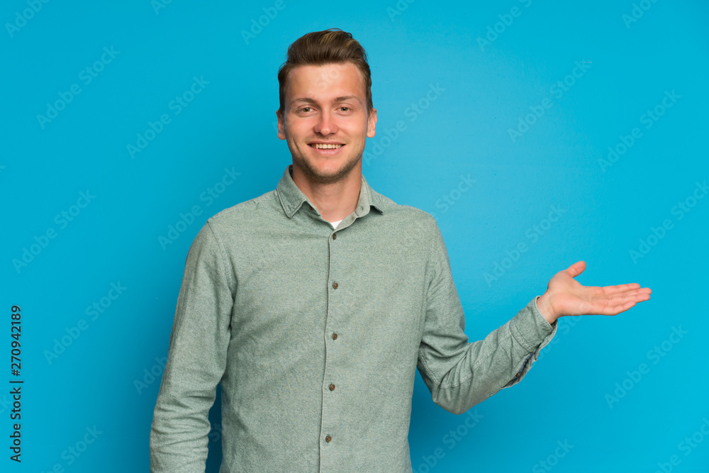 Blonde man over isolated blue wall holding copyspace imaginary on the palm to insert an ad Blonde man over isolated blue wall holding copyspace imaginary on the palm to insert an ad