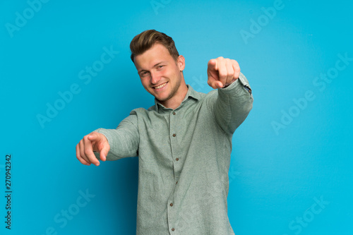 Blonde man over isolated blue wall points finger at you while smiling