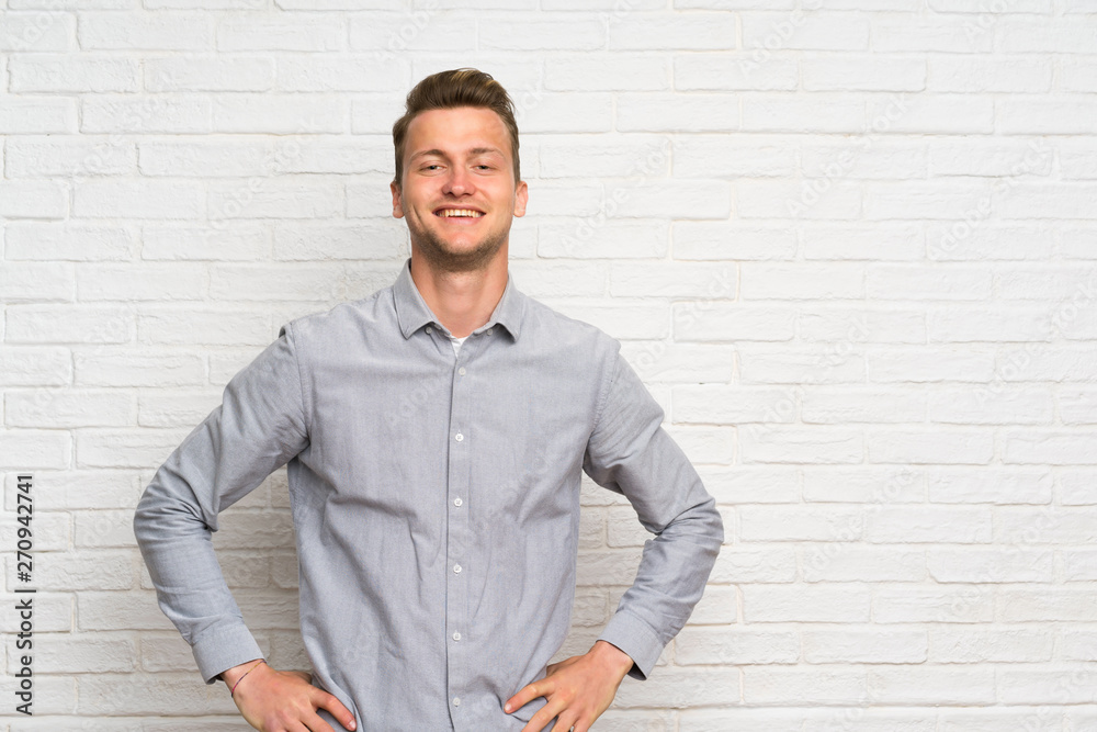 Blonde man over white brick wall posing with arms at hip and smiling Blonde man over white brick wall posing with arms at hip and smiling