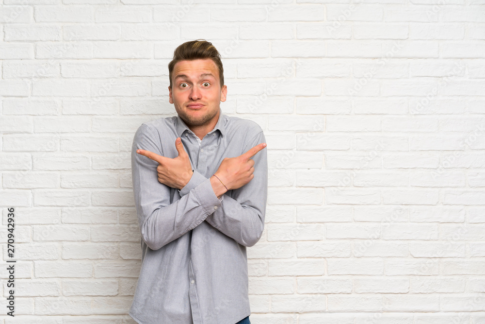 Blonde man over white brick wall pointing to the laterals having doubts Blonde man over white brick wall pointing to the laterals having doubts