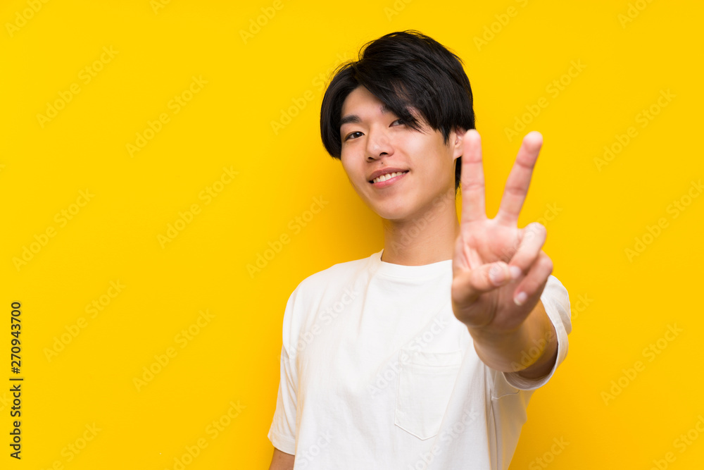 Asian man over isolated yellow wall smiling and showing victory sign Asian man over isolated yellow wall smiling and showing victory sign