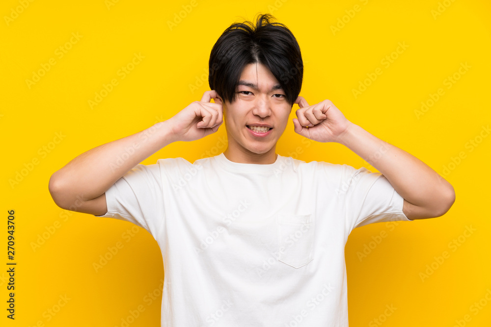 Asian man over isolated yellow wall frustrated and covering ears Asian man over isolated yellow wall frustrated and covering ears