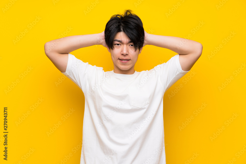 Asian man over isolated yellow wall frustrated and takes hands on head Asian man over isolated yellow wall frustrated and takes hands on head