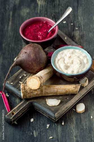 Two small round cups with freshly made seasoning, a horseradish root and beets on a dark wooden background. Selective focus.