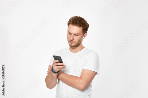 Studio shot of a bearded young man with wireless headphones holding looking at the screen of a mobile phone. © ianachyrva