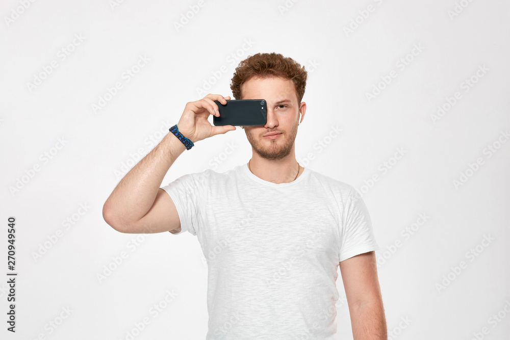 Studio shot of a bearded young man with wireless headphones holding mobile phone near the face and pretending to be looking through the phone camera. Studio shot of a bearded young man with wireless headphones holding mobile phone near the face and pretending to be looking through the phone camera.
