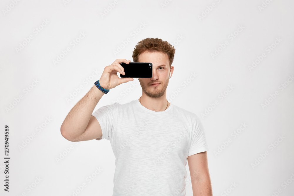 Studio shot of a bearded young man with wireless headphones holding mobile phone near the face and pretending to be looking through the phone camera. Studio shot of a bearded young man with wireless headphones holding mobile phone near the face and pretending to be looking through the phone camera.