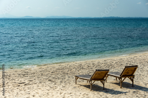Two Wooden chairs on the white sandy beach with little waves, blue and bright blue sky.