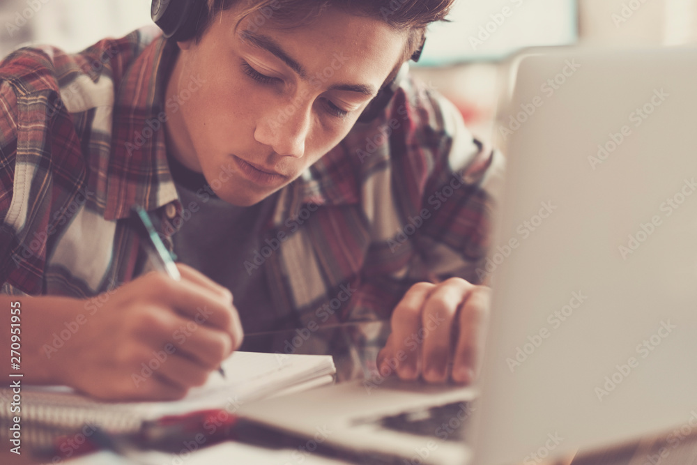 caucasian teenager indoor doing homework on the table at home - blonde guy writing and reading in his laptop or computer to get greats scores - portrait of boy with headphones caucasian teenager indoor doing homework on the table at home - blonde guy writing and reading in his laptop or computer to get greats scores - portrait of boy with headphones