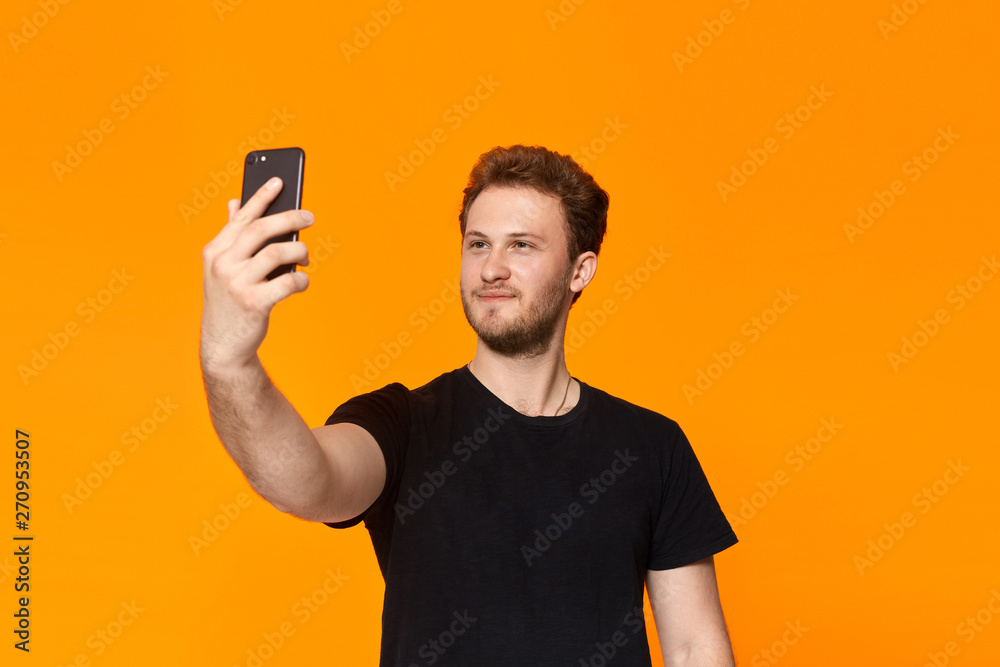 Studio shot of a smiling bearded young man making selfie with the smartphone while standing over orange background Studio shot of a smiling bearded young man making selfie with the smartphone while standing over orange background