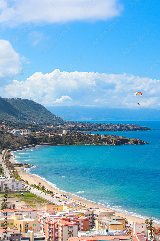 Paraglider flying over the amazing sea landscape by coastal Cefalu in Italian Sicily. Captured on a vertical photography with hills behind the beautiful bay