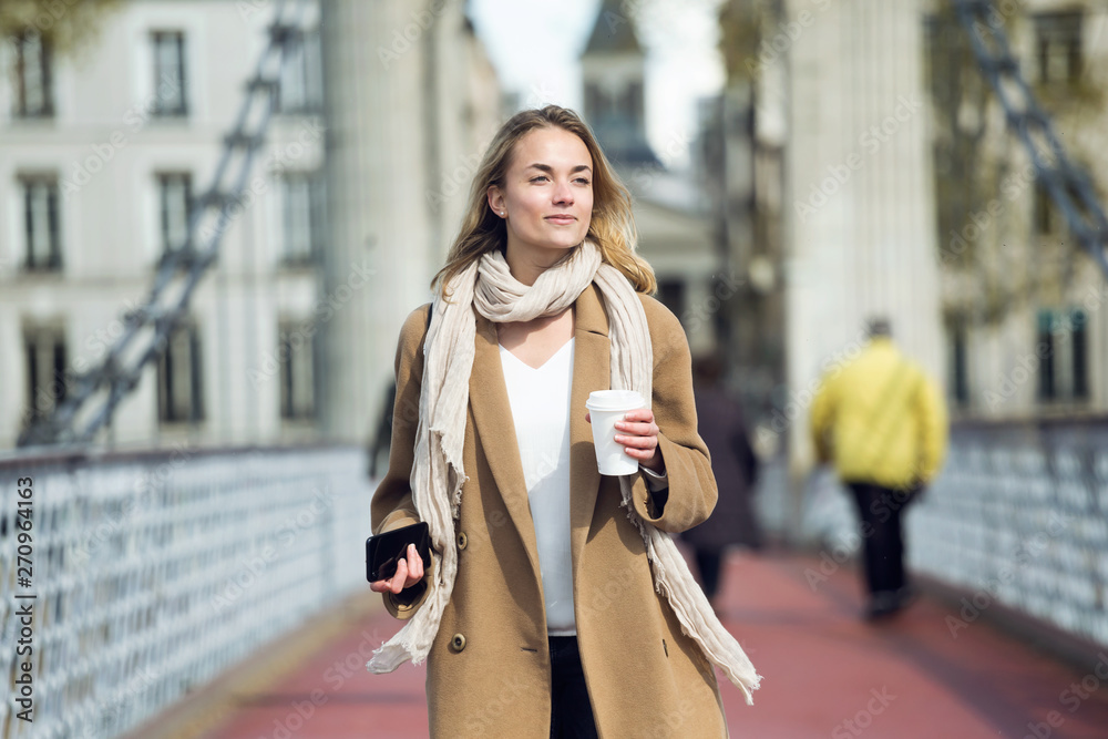 Pretty young woman holding cup of coffee and smartphone while walking in the street. Pretty young woman holding cup of coffee and smartphone while walking in the street.