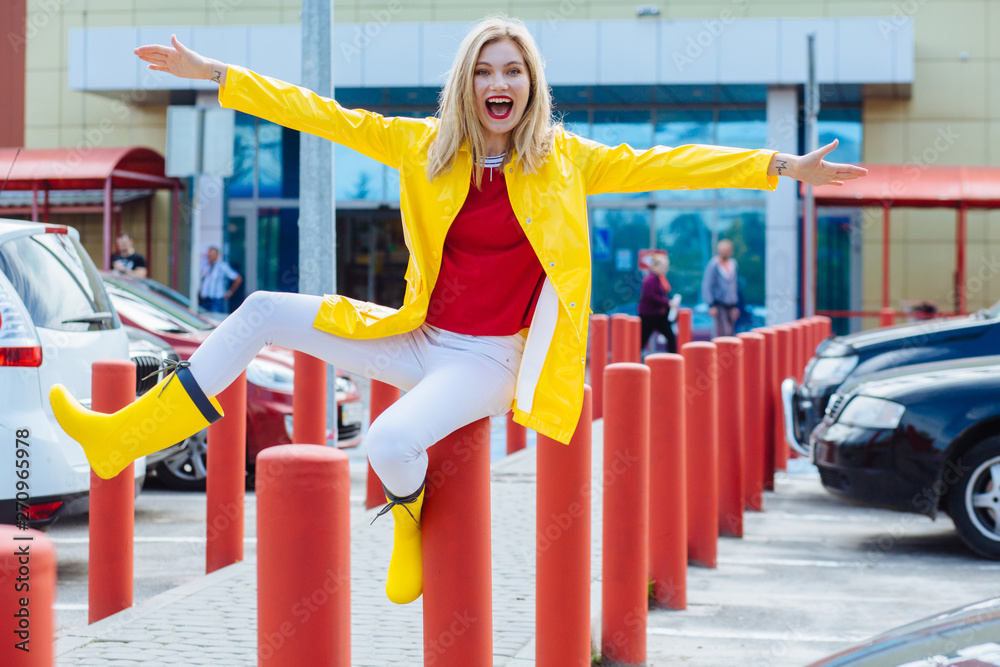 Full length portrait of a smiling blond teenager woman dressed in yellow raincoat red tishirt and rubber boots posing going crazy on parking over supermarket on background. Full length portrait of a smiling blond teenager woman dressed in yellow raincoat red tishirt and rubber boots posing going crazy on parking over supermarket on background.