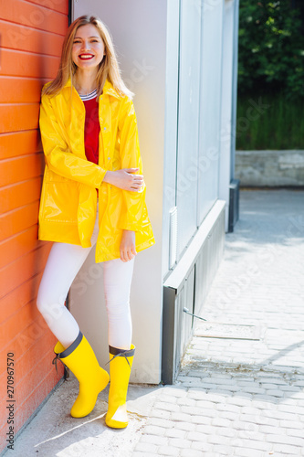 Full length portrait of a smiling blond teenager woman dressed in yellow  raincoat and rubber boots posing while standing on parking garage red door  on background. Outdoor lifestyle concept Stock-Foto | Adobe