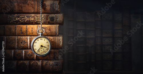 Vintage clock hanging on a chain on the background of old books. Old watch as a symbol of passing time. Concept on the theme of history, nostalgia, old age. Retro style.