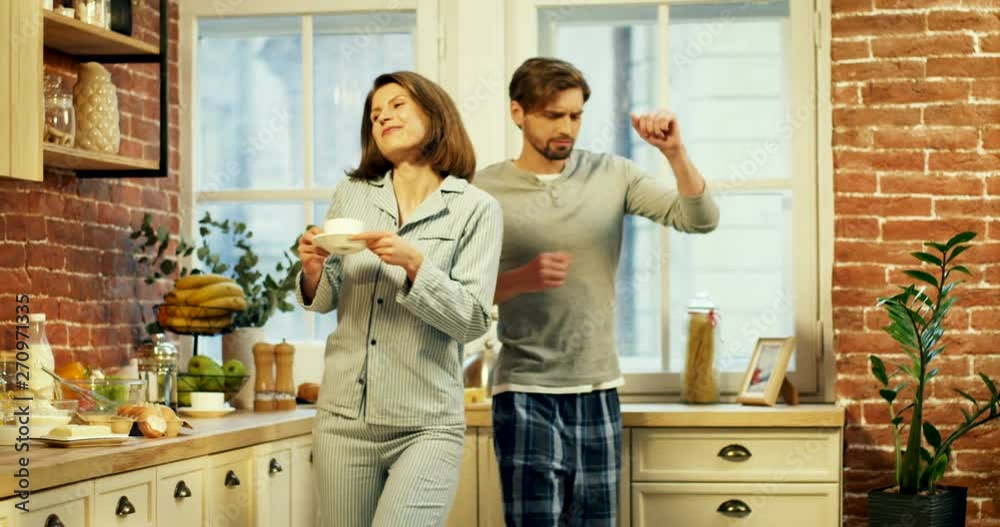 Hppy Caucasian young pretty woman and handsome man in pajamas dancing cheerfully in the kitchen in the morning and sipping coffee. Hppy Caucasian young pretty woman and handsome man in pajamas dancing cheerfully in the kitchen in the morning and sipping coffee.