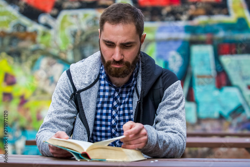 The guy with beard in plaid shirt sits at wooden table and reads the book against the background of the ornamented wall in graffiti.
