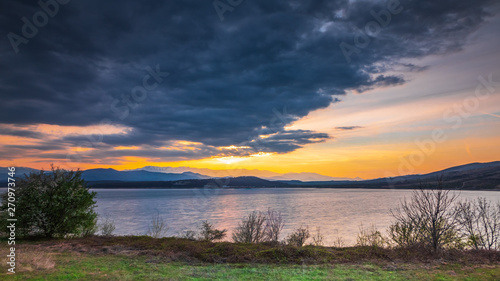A sunset on dark sky with dramatic clouds over a lake with distant hills in the background and a few trees and bushes in the foreground © Photo Shots