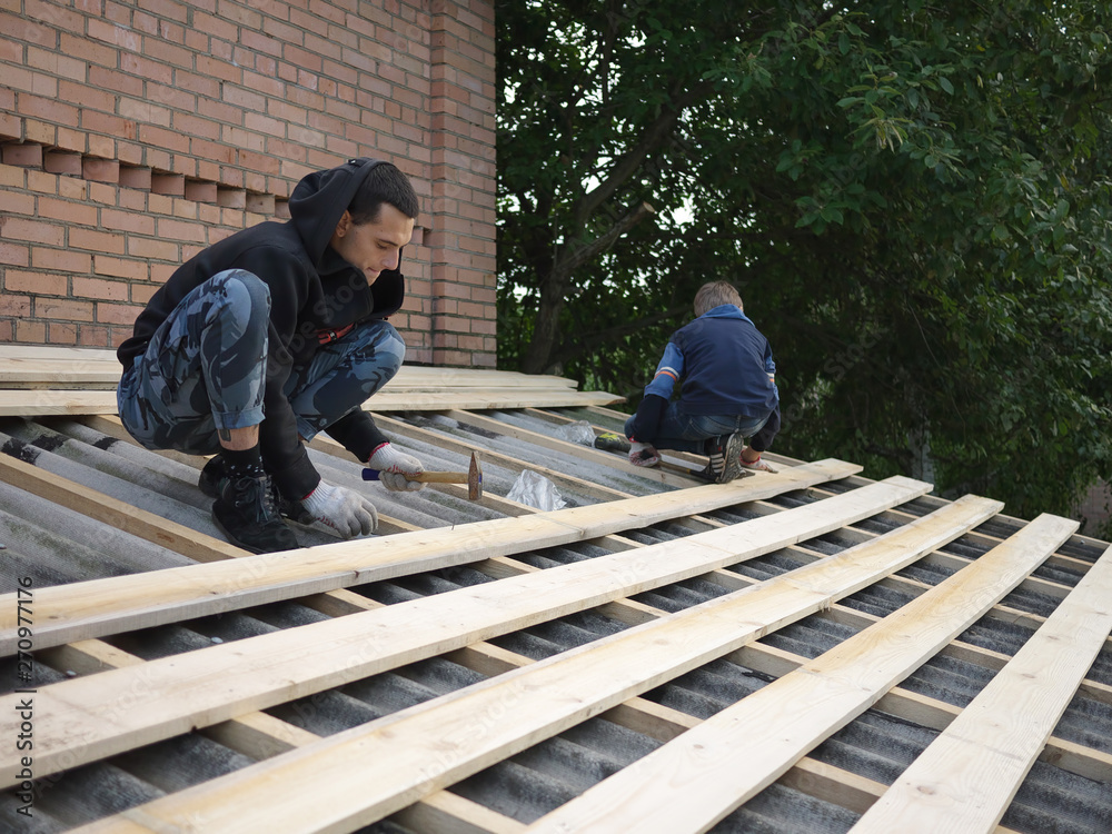 young caucasian man repairing the roof of a home; A worker replaces metal slate on the roof of a home young caucasian man repairing the roof of a home; A worker replaces metal slate on the roof of a home