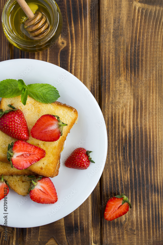 French toasts with strawberries and honey in the white plate on the brown wooden background.Top view.Copy space. French toasts with strawberries and honey in the white plate on the brown wooden background.Top view.Copy space.