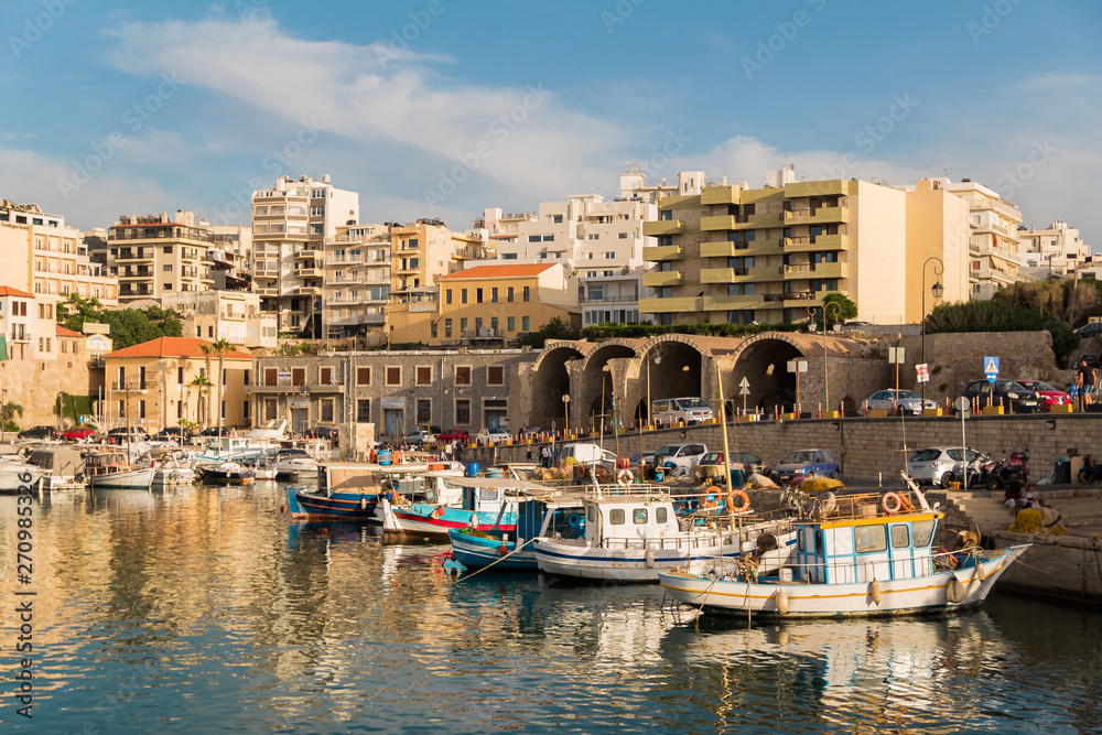 view of heraklion old port on a clear day. calm blue water boats floating and coastal road and buildings as background