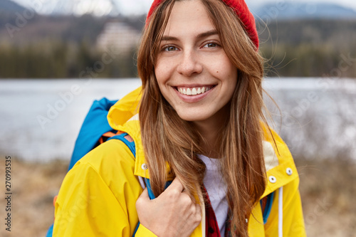 Cropped image of lovely cheerful European woman has broad tender smile, long straight hair, wears red hat, yellow anorak, has rucksack on back, poses over blurred nature background in open air © Wayhome Studio