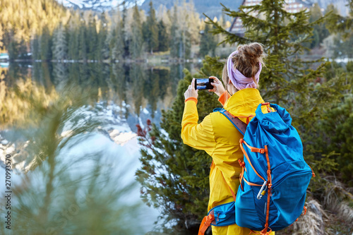 Freedom traveler takes pictures of scenic nature view, tries to capture beautiful lake with mountains and forest, stands back, carries big rucksack, explores hiking tours. People, recreation concept.
