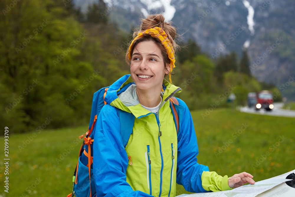 Naklejka premium Dreamy cheerful woman dressed in active wear, uses tourist map for finding right route, wears backpack, focused aside with glad expression, breathes fresh air in mountains, stands on green grass