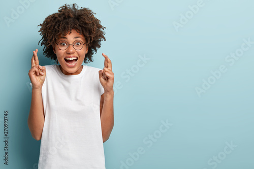 Studio shot of happy dark skinned woman crosses fingers for good luck, wears round glasses and casual white t shirt, believes dreams come true, models over blue background with empty space aside