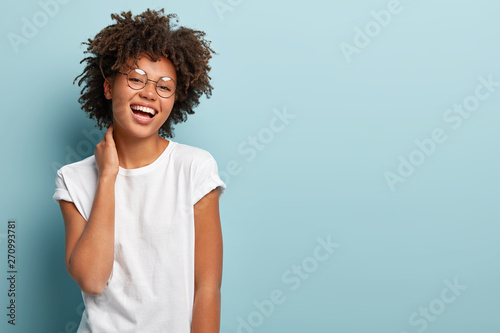 Portrait of happy carefree woman touches neck gently, demonstrates natural beauty, dressed in casual t shirt, smiles broadly, stands over blue background with empty space for your advertisement