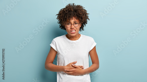 Dissatisfied young woman has food poisoning, keeps hands on belly, suffers from menstrual period cramp, feels discomfort in stomach, wears casual clothes, frowns face, isolated on blue background photo