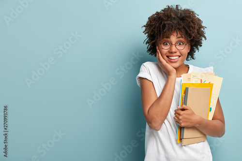 Pleasant looking Afro American woman holds notepads, papers, studies at college, glad to finish studying, keeps hand on cheek, wears white t shirt, isolated on blue background with blank space photo