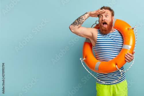 Surprised young holiday maker with red hair and beard, comes on beach with safety equipment as cannot swim, keeps hand on forehead, focused into distance, shocked to see drowning man in sea. photo