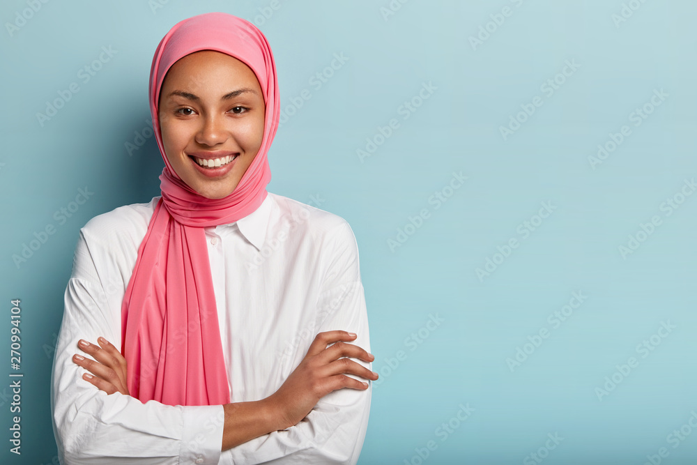 Studio shot of cheerful religious Muslim woman keeps arms folded, smiles broadly, has white teeth, expresses positive emotions, satisfied after praying, models over blue background with blank space Studio shot of cheerful religious Muslim woman keeps arms folded, smiles broadly, has white teeth, expresses positive emotions, satisfied after praying, models over blue background with blank space