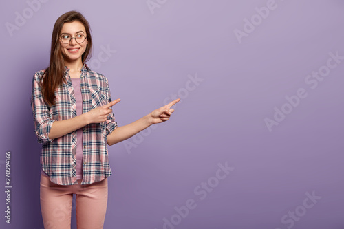 Gorgeous attractive woman shows product with discounts, has fun, promots item, has gentle smile, dressed in checkered shirt and trousers, isolated over purple wall, suggests going in this direction