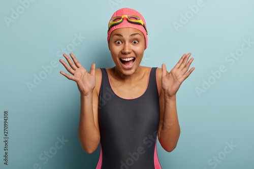 Candid shot of overemotive African American woman raises palms, has happy reaction, wears black swimsuit, special goggles, glad to spend recreation time on hobby, isolated over blue background. photo