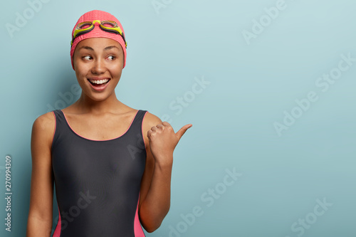 Studio shot of happy dark skinned female with charming smile, wears swimwear, points aside with thumb, shows direction to swim sport section, has glad expression, isolated over blue background