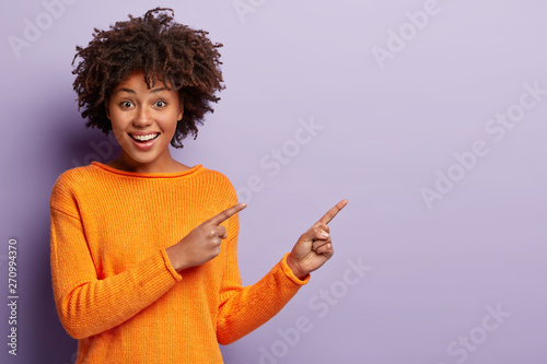Photo of delighted African American woman points away with both index fingers, promots awesome place for your advertising content, isolated over purple background, gives advice, shows copy space photo