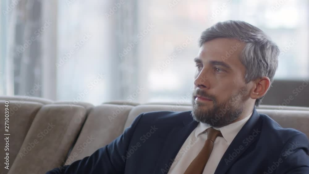 Close up of middle-aged grey-haired Caucasian businessman in suit sitting on sofa, relaxing watch strap, looking aside and then looking at camera Close up of middle-aged grey-haired Caucasian businessman in suit sitting on sofa, relaxing watch strap, looking aside and then looking at camera