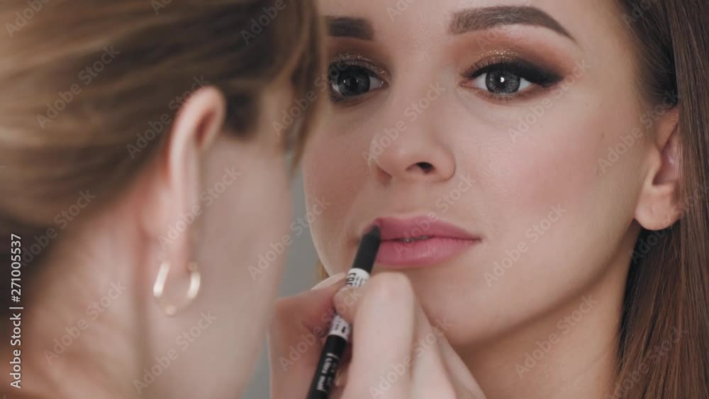 Closeup shot of cheerful young female makeup artist enjoying working with a beautiful young brown haired model, applying fresh trendy makeup on her lips with long eyelashes, perfect brows using pencil Closeup shot of cheerful young female makeup artist enjoying working with a beautiful young brown haired model, applying fresh trendy makeup on her lips with long eyelashes, perfect brows using pencil