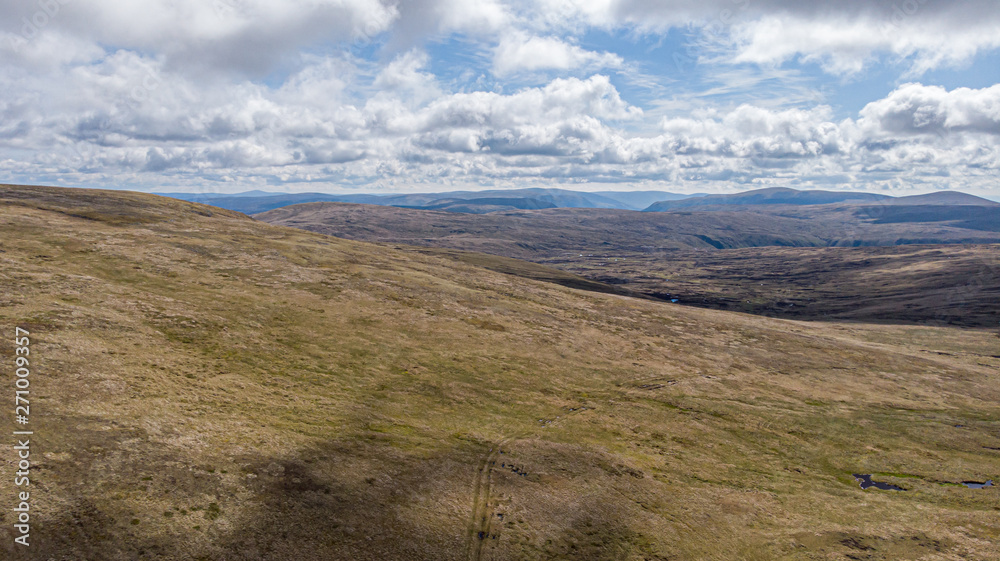 An aerial view of a Scottish moountain green heather slope with mountain range in the background under a majestic blue sky and white clouds