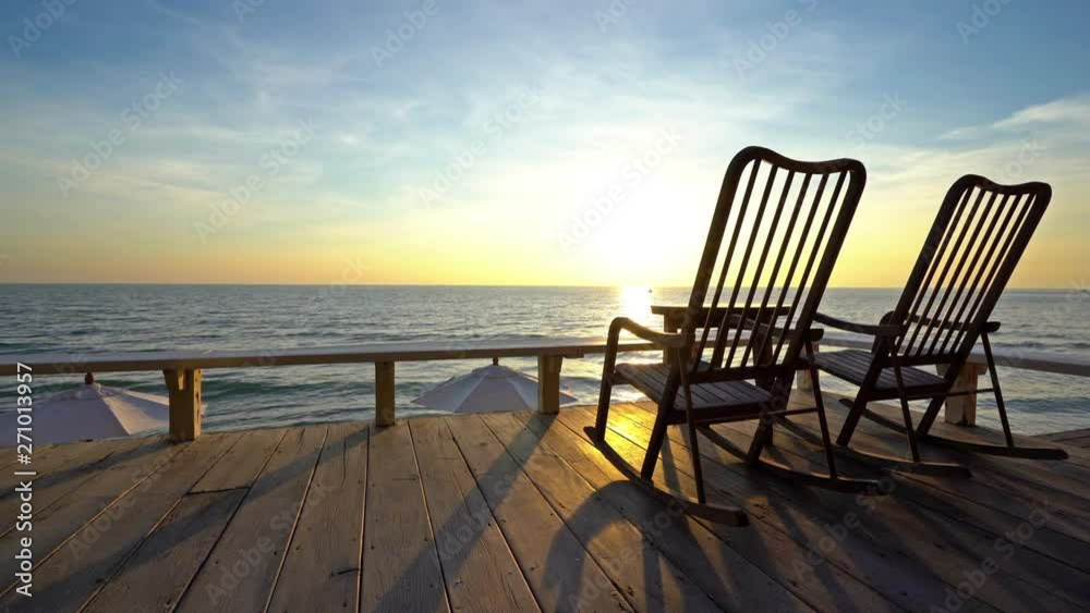 Locked off shot, two rocking chairs on wooden deck, golden sunset sea view background.