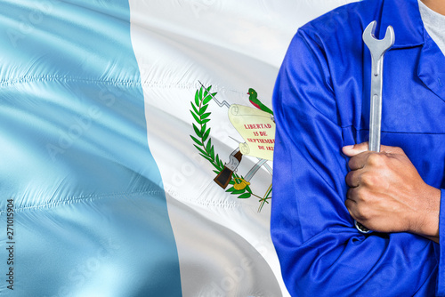 Guatemalan Mechanic in blue uniform is holding wrench against waving Guatemala flag background. Crossed arms technician.