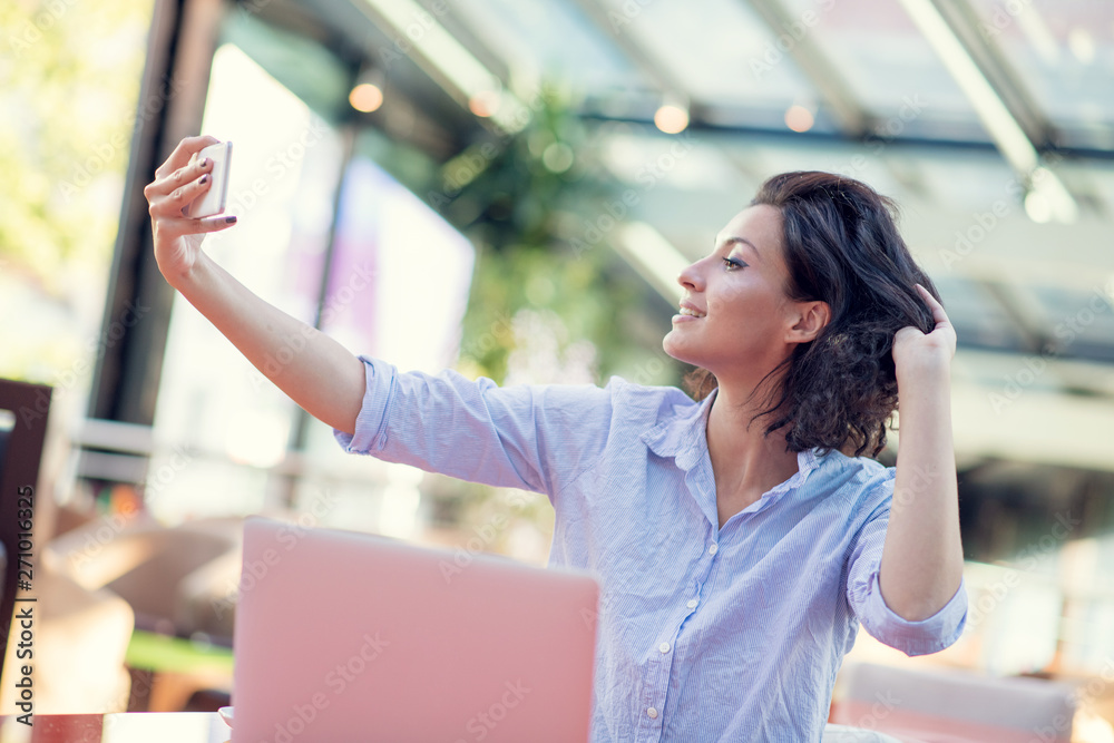 Portrait of a playful young girl taking selfie with mobile phone while sitting with laptop computer at a cafe outdoors